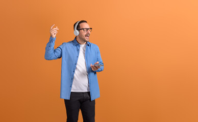 Young man with eyes closed singing song and listening music over headphones on orange background
