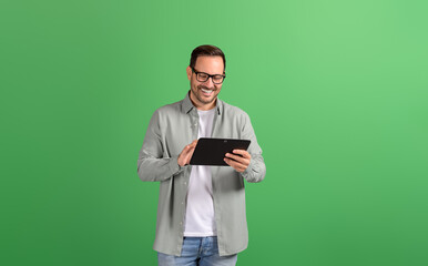 Smiling young businessman in eyeglasses reading e-mails over digital tablet on green background