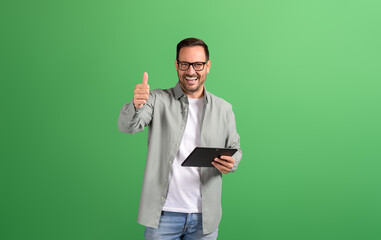 Portrait of happy handsome customer using touchpad and showing thumbs up sign on green background