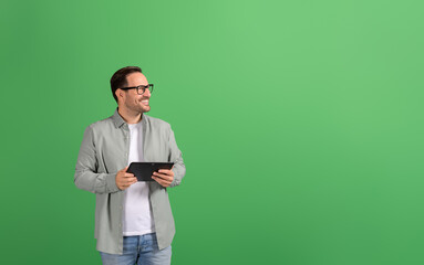 Successful businessman with digital tablet smiling and looking away thoughtfully on green background