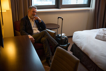 Smiling caucasian businessman with suitcase sitting in armchair in hotel room