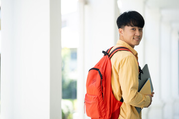 A college student stands in the hallway of his school, holding textbooks and smiling, ready for a...