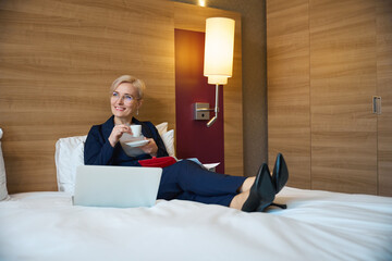 Smiling businesswoman drinking tea or coffee from cup and looking away on bed