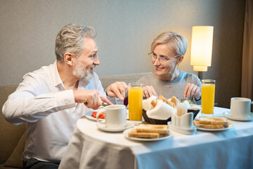 Fototapeta na wymiar Smiling mature couple eating breakfast and looking at each other in hotel room