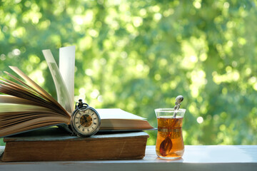 Glass tea cup, pocket watch and old books on wooden table in garden, green natural background. spring, summer season. atmosphere gentle image. Harmony, relax atmosphere