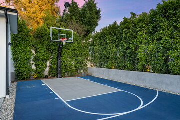 Outdoor basketball court by house with trees, bushes, and fence.