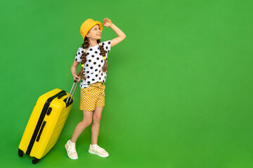 A little girl with a suitcase on wheels looks into the distance. A child in shorts and a hat is eating a vacation at the sea. Green isolated background. Copy space.