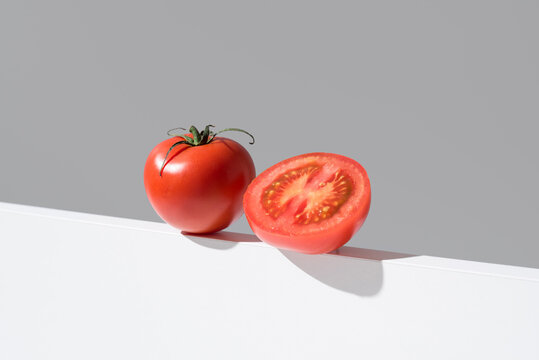 Un tomate maduro entero y cortado por la mitad sobre un soporte blanco y fondo gris