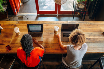 High angle view of two young women working on laptops side by side at a cafe counter.