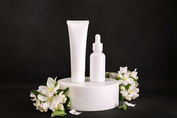 White unbranded cosmetic tubes on marble podium decorated with blooming apple twigs on black background close up.