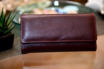 A maroon color ladies' purse made with premium quality leather, placed on the glass table and with...