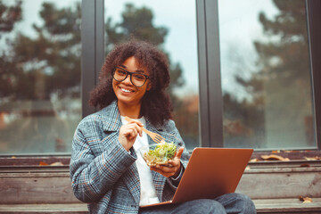 Happy african american woman eating salad, having healthy lunch while resting outdoors with laptop...