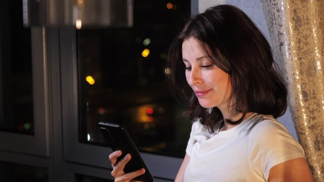 Young brunette is messaging with someone late at night, using her phone. She is surprised and smiles when she receives a message. Mid shot. 4k