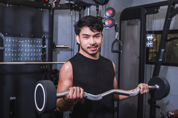 A fit asian man in a black tank top looks confidently while doing standing barbell curls at the gym. Using an EZ curl bar with fixed weight to build biceps.