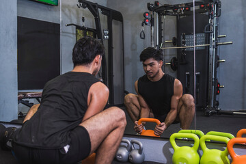 An asian man grabs an orange kettlebell from the floor to do a set of goblet squats. Training legs at the gym.