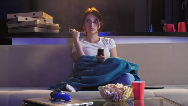 Young girl in glasses is sitting on the couch in the evening under a blanket, watching TV. She is smoking a vape and eating snacks while switching channels. Mid shot. 4k