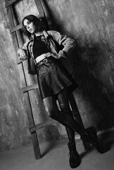 Indoor portrait of attractive grunge (rock) girl standing at wall. Informal model, dressed in a jean jacket, checkered shirt, leather skirt and holey tights. Black and white