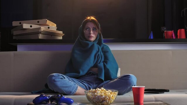 young girl is watching a horror movie with anxiety. She is tense, and she has wrapped herself in a blanket to make it less scary. Mid shot. 4k