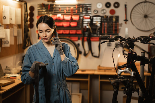 Female mechanic takes order by mobile phone standing in bicycle workshop near bike. Mechanic talking on mobile phone after fixing bike