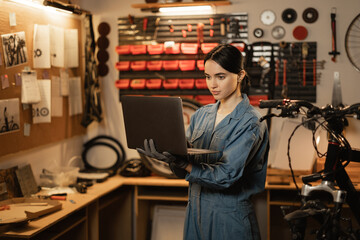 Brunette woman in casual clothes using a laptop and smiling while standing in the bicycle repair shop
