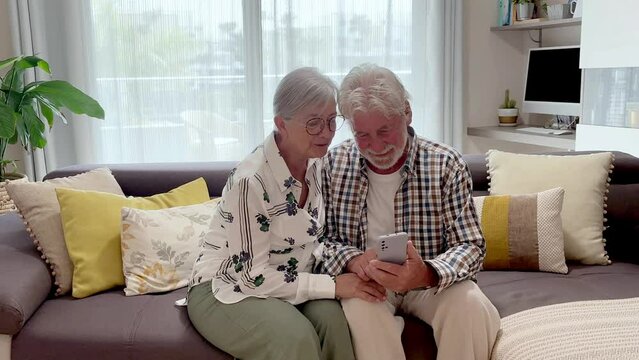 Smiling senior couple sitting on home sofa using smart phone enjoying technology, elderly 70 years old caucasian people sitting together in living room following social media, serene retirement lifest