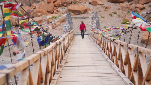 Handheld shot of wooden bridge above the Tsarap Chu river with prayer flags waving on the side due to the strong winds with defocused man walking on the bridge during trek to Phugtal Gompa.