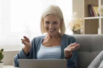 Overjoyed older female share news during on-line talk to family, resting on sofa looking at laptop...