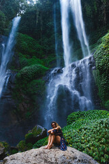 Fototapeta na wymiar Young Women Sitting On The Rock Near The Sekumpul Waterfall, Bali, Indonesia