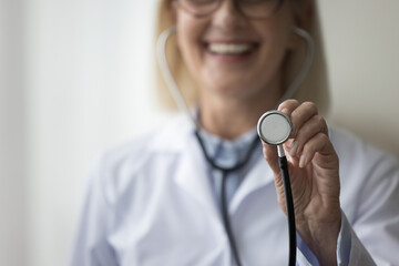 Close up of smiling mature woman cardiologist showing to camera stethoscope. Assessing patient health, listen internal body sounds, check health condition of heart or lungs, measure of blood pressure - obrazy, fototapety, plakaty