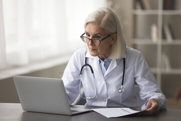 Mature woman therapist working on laptop, sit at desk with papers, prepare treatment plan, check patient medical records, make research and clinical guidelines, reviewing data, do administrative tasks