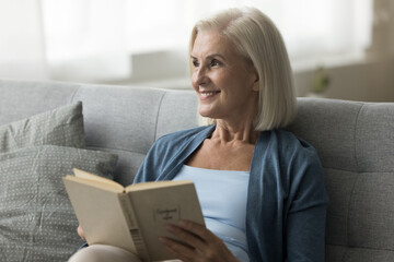 Cheerful senior 60s woman sit on cozy sofa read interesting book, looking away, enjoy literature...