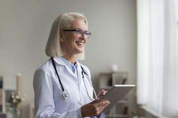 Smiling mature cardiologist using tablet at workplace. Modern device usage to order diagnostic tests, prescriptions electronically, using medical resources, clinical guidelines, and educational videos - obrazy, fototapety, plakaty