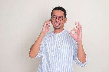 Fototapeta premium Adult Asian man smiling and give OK finger sign while answering a phone call