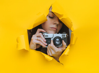 A young paparazzi girl with a rare SLR camera looks out from her hiding place and carefully watches what is happening. Yellow paper hole. Tabloid press. Looking for a subject for stock photos. - obrazy, fototapety, plakaty