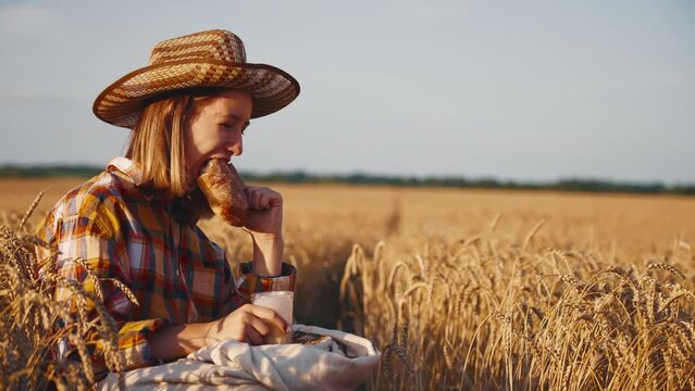 Young girl farmer in sun hat eats loaf of bread biting it drinking milk sits on wheat field with grain bag on knees. Harvesting, summer on farm, farmland, agribusiness, woman worker has break.