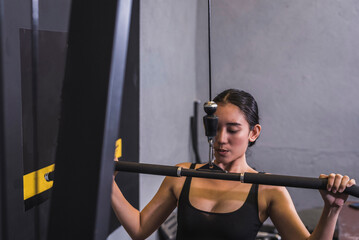 A young asian woman performs wide grip straight lat pulldowns. Weight training targeting back muscles and upper body. Shot from behind.