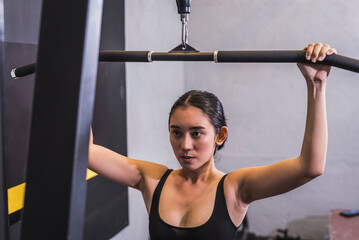 A determined young asian woman performs wide grip straight lat pulldowns. Weight training targeting back muscles and upper body.