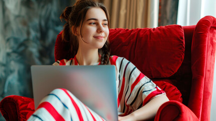 A beautiful young woman in a striped tunic with a laptop on her lap is sitting in a red armchair. - obrazy, fototapety, plakaty