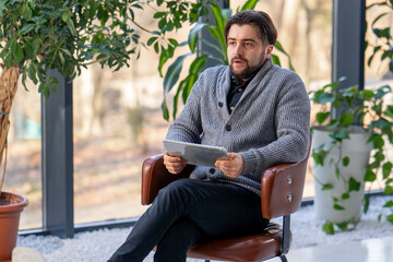 Fototapeta premium In a white hall young guy office worker on a brown chair works on a tablet