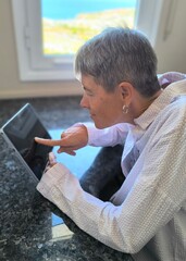 Adult woman with short gray hair, sitting at the desk with the tablet in her hand, looking with her...