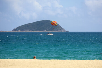 Sea beach with sand, view to motorboat with parachute and tourists sailing on mountain island background. Background for holidays on a paradise nature
