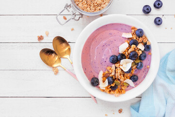 Healthy blueberry and coconut smoothie bowl with granola. Above view table scene on a white wood background.