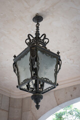 Detail of an old and ornate wrought iron lamp hanging from the ceiling. It is square in shape and...