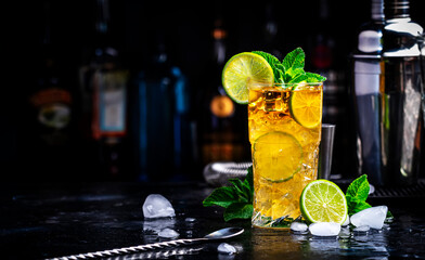 Dark and stormy alcoholic cocktail long drink with dark rum, ginger ale, lime and ice with bottles, black bar counter background