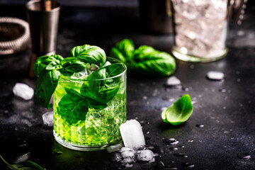 Green summer alcoholic cocktail drink with dry gin, sugar syrup, lemon, green basil and ice, dark bar counter background