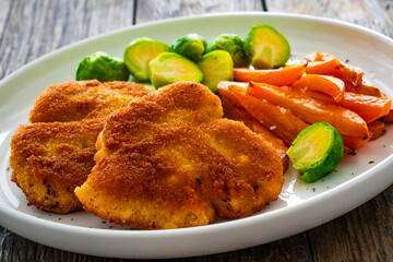 Crispy breaded fried pork loin chops with sweet potato fries and boiled Brussels sprouts on wooden table
