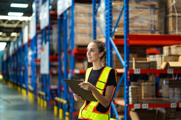 A woman wearing a safety vest and holding a clipboard is standing in a warehouse. She is looking at...