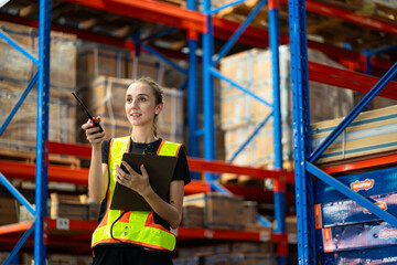 A woman wearing a safety vest and holding a clipboard is standing in a warehouse. She is smiling...