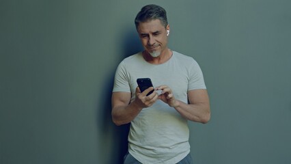 Casual male in 50s with earphones using a mobile phone. Portrait of happy man smiling. Blank copy space for text on grey wall background.