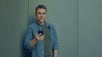 Portrait of a smiling middle-aged man holding a phone. Happy, confident mid adult male in casual. Blank copy space on a gray wall background.
